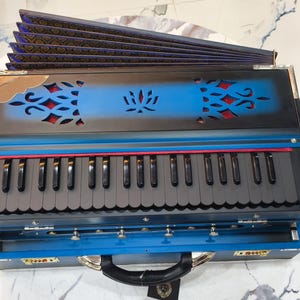 May include: A blue and black harmonium with a keyboard and bellows. The instrument has decorative cutouts and a handle. The keyboard has black and white keys, and the bellows are folded. The harmonium is on a white surface.