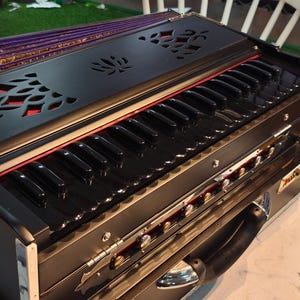 Könnte beinhalten: Ein schwarzes und silbernes Harmonium mit einer Tastatur und dekorativen Ausschnitten. Das Instrument hat eine lila und goldene Oberseite und einen Griff. Das Harmonium ist ein Musikinstrument, das in der indischen klassischen Musik verwendet wird.