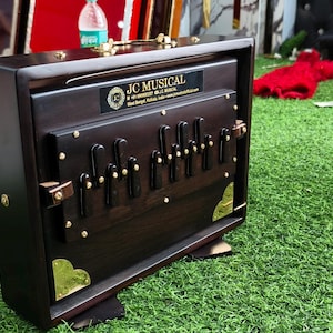 May include: A dark brown wooden harmonium with gold-colored accents and the text "JC MUSICAL". The instrument has a series of black keys and is set on a green surface.