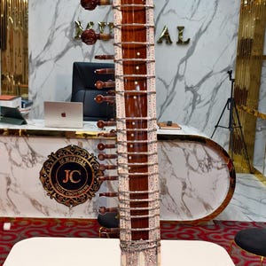 Può includere: Un sitar con un manico in legno lucido marrone scuro e una grande cassa di risonanza arrotondata con intagli floreali complessi. Lo strumento presenta numerose corde metalliche, tasti argentati e motivi decorativi a intarsio bianco.