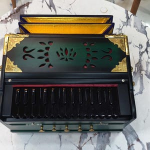 May include: A green and black harmonium with gold detailing, set on a marble-effect surface. The instrument has a keyboard, decorative cutouts, and a red interior. The top section has three panels with gold trim. A traditional musical instrument.