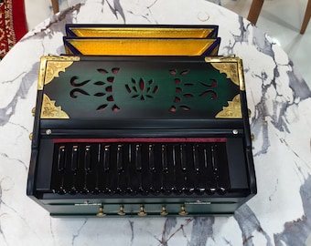 May include: A green and black harmonium with gold accents, placed on a marble-patterned surface. The instrument features a keyboard, decorative cutouts, and a red interior. The top section has three panels with gold trim. A traditional musical instrument.