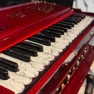 May include: A close-up of a red harmonium with black and white keys. The wooden instrument has intricate carvings on the top and is positioned at an angle, showcasing the keyboard and the polished finish.