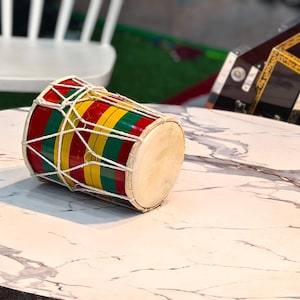 Op de afbeelding: Een kleine, ronde trommel met rode, gele en groene strepen. De trommel is gemaakt van hout en heeft een leren vel. Hij staat op een witte marmeren tafel.