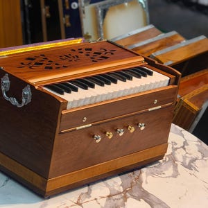 May include: A wooden harmonium with a keyboard and decorative carvings on the top. The instrument has a handle and several knobs. Other harmoniums are visible in the background. The harmonium is on a marble-patterned surface.