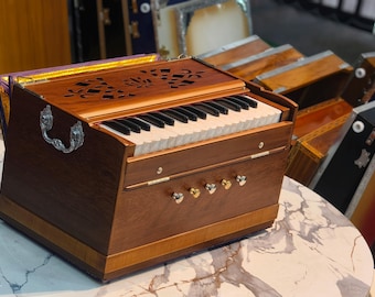 Pode incluir: Um harmônio de madeira com teclado e entalhes decorativos na parte superior. O instrumento tem uma alça e vários botões. Outros harmônios são visíveis ao fundo. O harmônio está sobre uma superfície com padrão de mármore.
