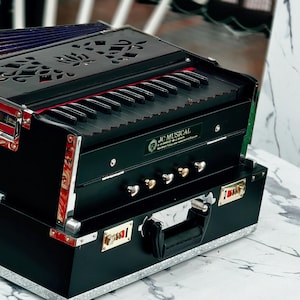 May include: A black and gold harmonium with a keyboard and decorative top. The instrument is in a black case with silver trim and a handle. The harmonium is on a white marble-patterned surface.