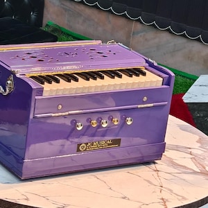 May include: A purple harmonium with black and white keys sits on a round marble table. The instrument has silver hardware and the text "J.L. MUSICAL" on the front. A white chair is to the left.