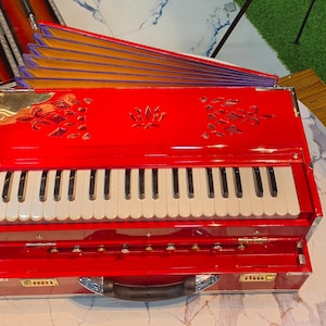 May include: A red harmonium with white keys and a handle. The musical instrument has decorative cutouts on the top and is sitting on a white surface. Other instruments and cases are in the background.