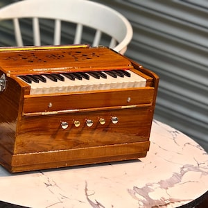 Könnte beinhalten: Ein hölzernes Harmonium mit Tastatur und dekorativen Schnitzereien. Das Instrument ist braun mit silberfarbener Hardware und steht auf einem runden Tisch mit Marmormuster. Gitarren sind im Hintergrund zu sehen.