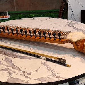 May include: A wooden Indian stringed instrument, a sitar, with a long neck and multiple strings, resting on a marble-patterned table. A bow lies beside it. The sitar has a light brown body and dark tuning pegs.