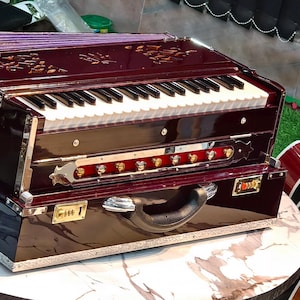 May include: A dark red harmonium with a keyboard and a case. The harmonium has a decorative design on the top and a handle on the case.