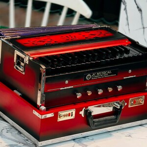 May include: A red and black harmonium with a keyboard and decorative accents. The instrument is in a red carrying case. The text "JC MUSICAL" is visible on the front of the harmonium.