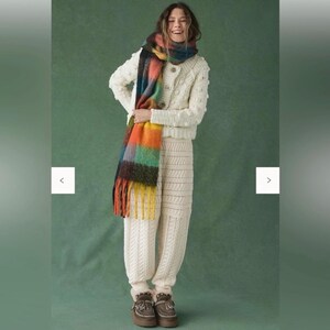May include: A woman in a cream-coloured knitted cardigan and matching knitted trousers. A colourful scarf with orange, yellow, green, and blue hues is draped around the neck. The outfit is paired with brown boots.