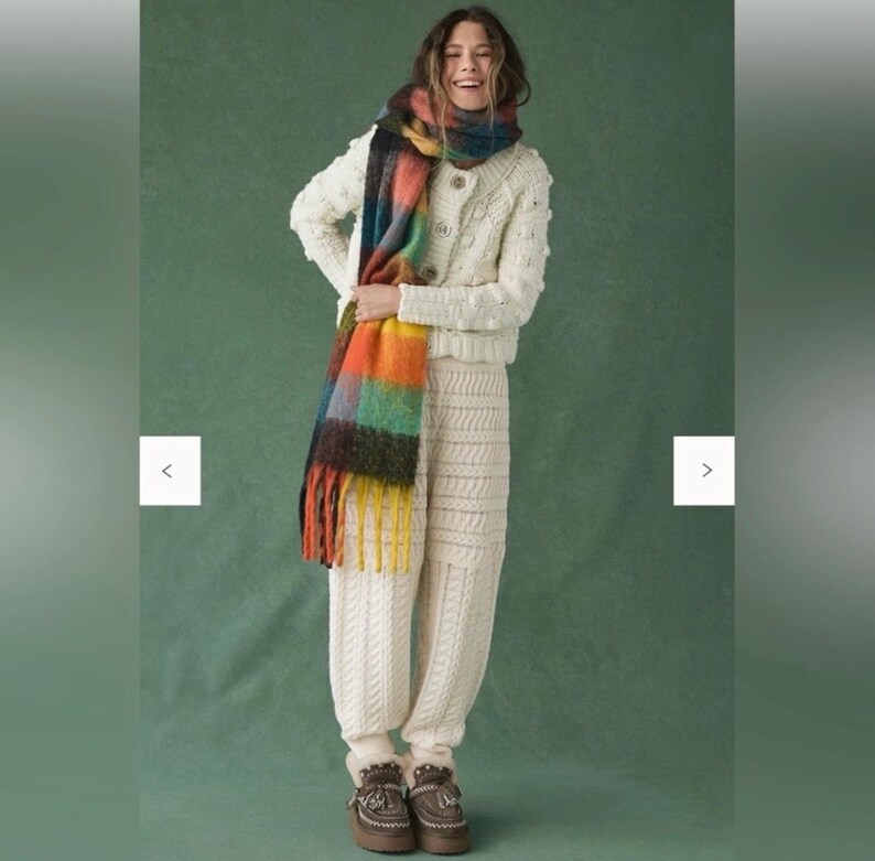 May include: A woman in a cream-coloured knitted cardigan and matching knitted trousers. A colourful scarf with orange, yellow, green, and blue hues is draped around the neck. The outfit is paired with brown boots.