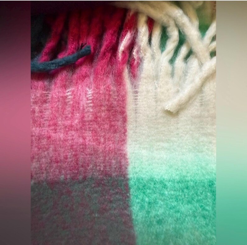 May include: Close-up of a colourful woven blanket with a fringed edge. The blanket features a colour block design with sections of pink, white, green, and dark teal. The texture appears soft and fuzzy.