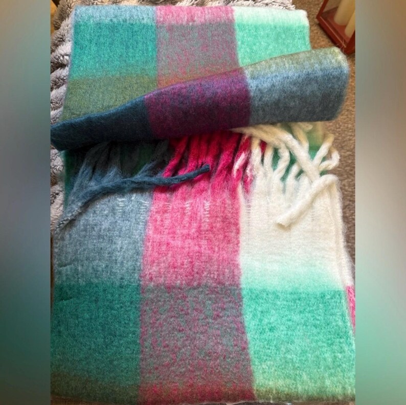 May include: A colourful blanket with a plaid design. The blanket has squares of teal, pink, purple, and grey. The blanket is rolled up and has a fringe of white and teal tassels.