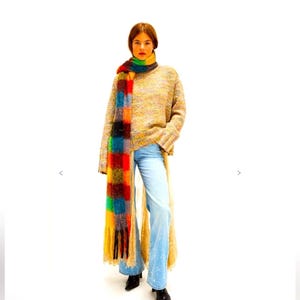 May include: A person wearing a multi-coloured scarf, a beige sweater, and light blue jeans. The scarf has a patchwork design with red, orange, yellow, green, and blue squares. The sweater is a loose knit, and the jeans are flared.