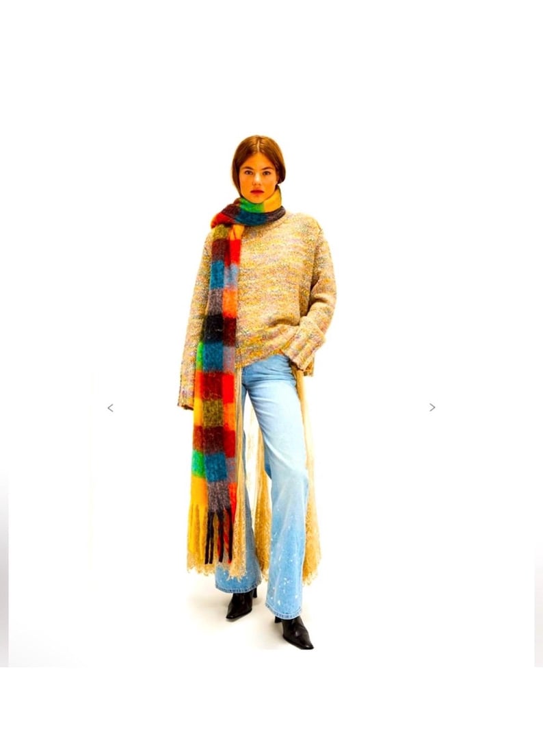 May include: A person wearing a multi-coloured scarf, a beige sweater, and light blue jeans. The scarf has a patchwork design with red, orange, yellow, green, and blue squares. The sweater is a loose knit, and the jeans are flared.