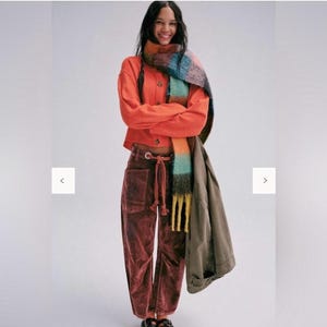 May include: A woman is wearing an orange cardigan, burgundy velvet trousers with a drawstring waist, and a colourful scarf with fringe. A green jacket is draped over the shoulder. The model is smiling.