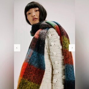 May include: A vibrant, oversized scarf featuring a plaid design in red, blue, green, and black. The scarf is draped over a cream-coloured sweater with a textured bobble stitch pattern. The scarf appears to be made of a soft, fuzzy material.