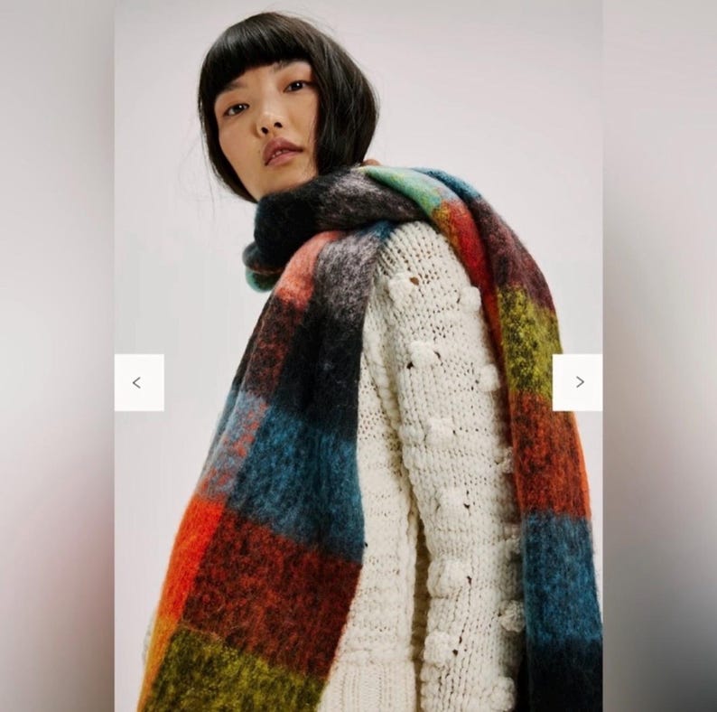 May include: A vibrant, oversized scarf featuring a plaid design in red, blue, green, and black. The scarf is draped over a cream-coloured sweater with a textured bobble stitch pattern. The scarf appears to be made of a soft, fuzzy material.