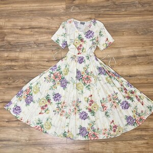 May include: A white dress with a floral print featuring purple, pink, and green flowers. The dress has a pleated skirt and short sleeves.