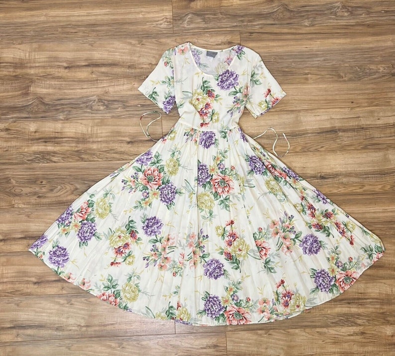May include: A white dress with a floral print featuring purple, pink, and green flowers. The dress has a pleated skirt and short sleeves.