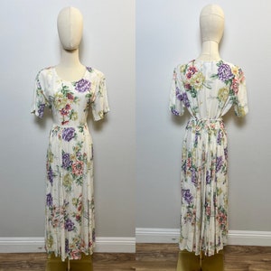 May include: A white maxi dress with a floral print featuring pink, purple, and green flowers. The dress has a round neckline, short sleeves, and a pleated skirt.