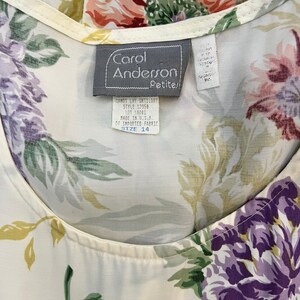 May include: A white blouse with a floral print in shades of green, purple, pink, and red. The blouse has a scoop neck and a label that reads "Carol Anderson Petites!" with the text "Banos Lay Untilory Style 12350 Lot 16001 Made in U.S.A. of Imported Fabric Size 14."