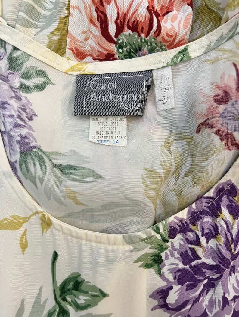 May include: A white blouse with a floral print in shades of green, purple, pink, and red. The blouse has a scoop neck and a label that reads "Carol Anderson Petites!" with the text "Banos Lay Untilory Style 12350 Lot 16001 Made in U.S.A. of Imported Fabric Size 14."