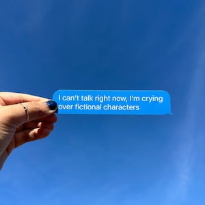 May include: A hand holds up a blue text message bubble against a bright blue sky. The text reads, "I can't talk right now, I'm crying over fictional characters."