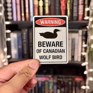 Op de afbeelding: Een rechthoekige sticker met een zwart silhouet van een vogel en de tekst "WARNING" in een rode ovaal. Daaronder staat de tekst "BEWARE OF CANADIAN WOLF BIRD" in vette, zwarte letters. De sticker wordt voor een wazige boekenplank gehouden.