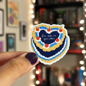 May include: A blue and yellow heart-shaped cake with white frosting and red cherries. The cake has the text "fine, make me your villain" written on it.