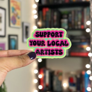 May include: A sticker with a multi-colored, retro-style font that reads "Support Your Local Artists".