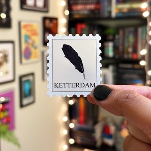 white border (that mimics a postage stamp shape) around an off-white rectangle. "Ketterdam" written in black on the bottom. A black feather is placed in the center
