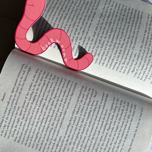 May include: A pink bookmark shaped like a worm with the words "Bookworm" written on it. The bookmark is placed on an open book with text on the pages.