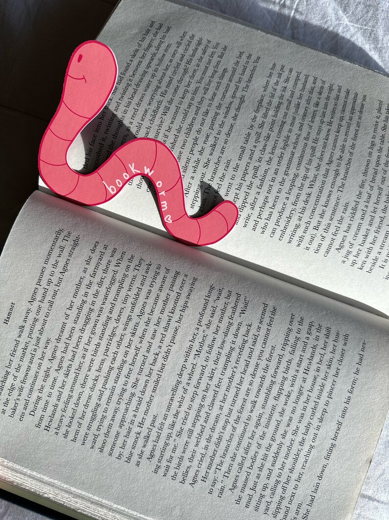 May include: A pink bookmark shaped like a worm with the words "Bookworm" written on it. The bookmark is placed on an open book with text on the pages.