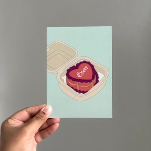 Puede incluir: Ilustración de un pastel en forma de corazón con la palabra "Books" encima, presentado en un recipiente para llevar. El pastel es rojo y morado, sobre una tarjeta azul claro.
