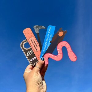 May include: A set of four colorful bookmarks featuring a pink worm, a blue text bubble, a black and white crow, and a pink and white band-aid. The bookmarks have text that reads "I'm reading", "Can't talk right now, I'm crying", "Emotional characters", and "Bookmark".