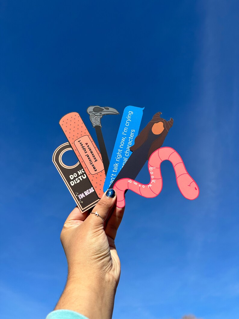 May include: A set of four colorful bookmarks featuring a pink worm, a blue text bubble, a black and white crow, and a pink and white band-aid. The bookmarks have text that reads "I'm reading", "Can't talk right now, I'm crying", "Emotional characters", and "Bookmark".