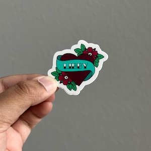 May include: A sticker featuring a heart-shaped design with a banner that reads "LIBBY". The heart is burgundy with a teal banner, surrounded by red flowers and green leaves. The sticker has a white border.
