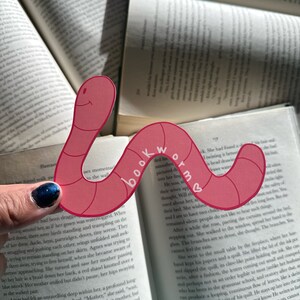 May include: A pink and red bookmark shaped like a worm with the word "bookworm" written on it. The bookmark is being used to mark a page in a book.