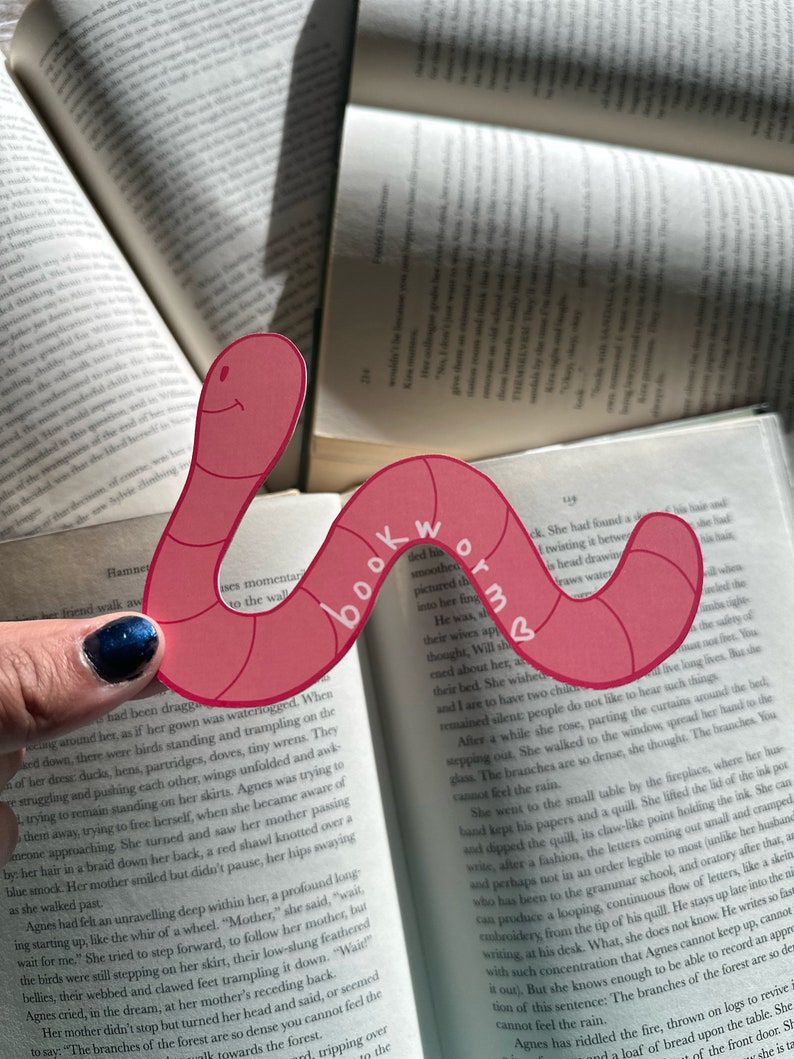 May include: A pink and red bookmark shaped like a worm with the word "bookworm" written on it. The bookmark is being used to mark a page in a book.