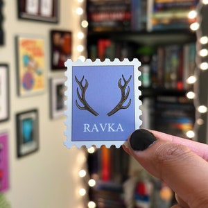 May include: A purple and white sticker with a stamp design. The sticker features a pair of antlers and the word "RAVKA" in black text.