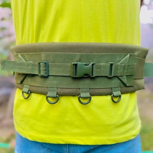 May include: A green tactical belt with multiple D-rings and a buckle closure. The belt is worn around a person's waist.