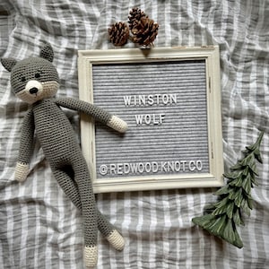 May include: A handmade gray crochet wolf doll with white accents on the paws and snout. The doll is lying next to a framed letter board that reads "WINSTON WOLF" and the text "@REDWOODKNOTCO". Decorative pinecones and a small green tree are also present.