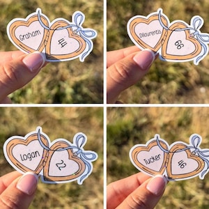 May include: A set of four heart-shaped stickers featuring a light orange outline and a pale pink center. Each sticker displays a name and a two-digit number, including Graham 44, Dilaurentis 66, Logan 22, and Tucker 46, accented with a light blue bow.