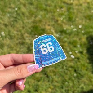 Pu&ograve; includere: Un adesivo blu a forma di maglia da hockey con il nome "Di Laurentis" e il numero "66" in bianco. La maglia ha un design glitterato e colorato. L'adesivo &egrave; tenuto su uno sfondo di erba verde.