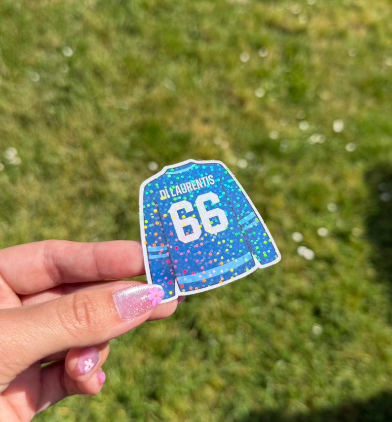 Pu&ograve; includere: Un adesivo blu a forma di maglia da hockey con il nome "Di Laurentis" e il numero "66" in bianco. La maglia ha un design glitterato e colorato. L'adesivo &egrave; tenuto su uno sfondo di erba verde.